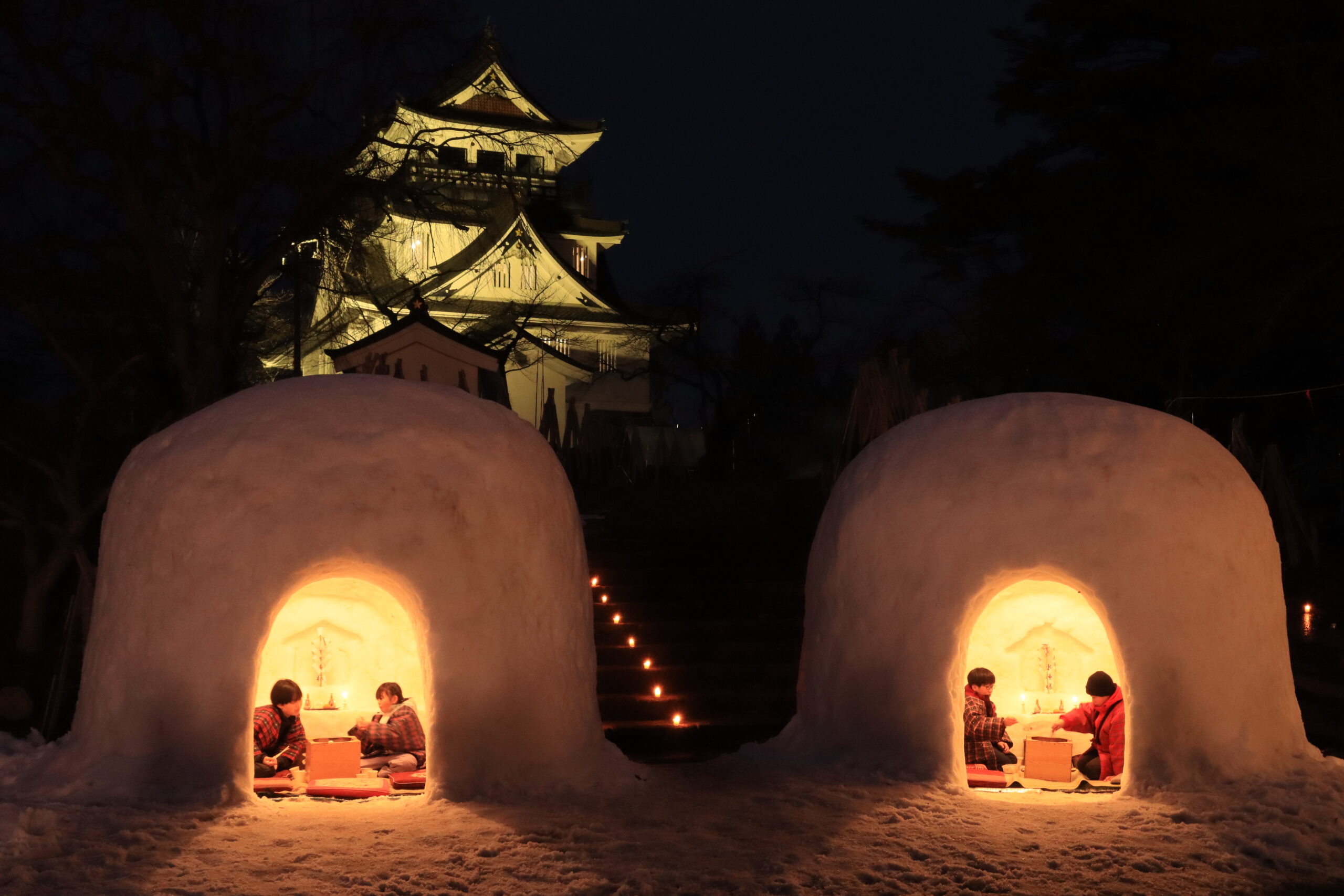 Warmly lit kamakura snow huts in Yokote, Akita, with visitors sitting inside while a traditional Japanese castle glows in the background.